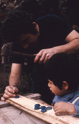 Hombre junto a niño pequeño clavando tapas metálicas en una tabla